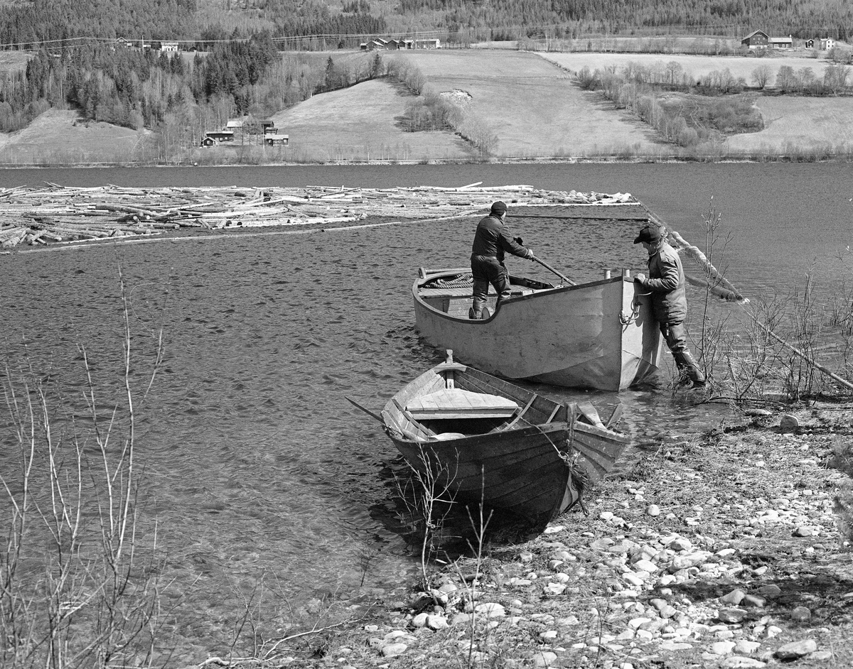 Båter og tømmerbom ved Lomnessjøens vestre bredd, antakelig i nærheten av elva Ottas utløp ved Otnes, der Glomma fellesfløtingsforening hadde ei koie for mannskapene sine (ikke synlig på dette fotografiet).   Ved strandlinja i forgrunnen lå det en robåt av tre og en varpebåt av stål.  Den sistnevnte båttypen ble brukt til å slepe tømmer i bommer sørover mot Kvernnesodden, der bommene ble åpnet.  I varpebåten står Harald Hansen (1926-1999), mens Ola Stubsveen (1924-1998) står ved standlinja. I bakgrunnen ser vi innsjøens østside med innmarka Lomnes-gardene og noe av den ovenforliggende skogen.