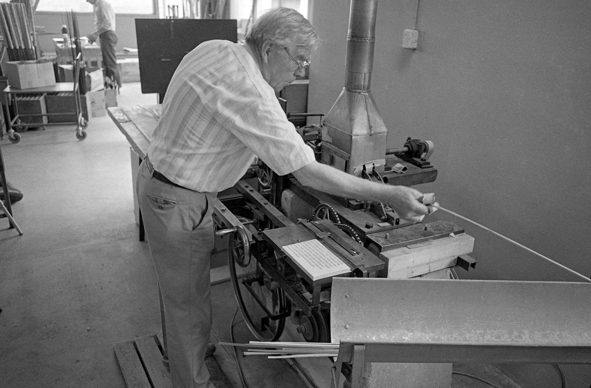 Industrigründeren Asbjørn Hørgård (1910-2001), fotografert i pruduksjonslokalene til bedriften han bygde opp i Trondheim.  Hørgård var særlig kjent for sine laminerte fiskestenger, såkalte splitcanestenger.  Veden i stengene var fra en bambusart som ble importert fra Østen.  Hørgård brukte også begrepet «tonkin», som antakelig henspilte på den sørøstasiatiske regionen råstoffet ble hentet fra.  Bambusen ble splittet i flere seksjoner (spildrer) og deretter limt sammen igjen.  Hørgård skrev følgende: «Den konstruksjonen som ble den seirende var den hvor spildrene ble høvlet i 60 graders vinkel og limt sammen til et enkeltbygget, sekskantet stangemne.  I grovere laksestenger ble det brukt dobbelt byggede stenger, dette på grunn av bambusspildrenes begrensede tykkelse.  Som konkurransestenger ble enhåndsstenger vesentlig hulbygget for å lette vekten og øke aksjonen».  Bambusstavene ble importert i 12 fots lengder og lagret i lang tid før bearbeidinga startet.  I fiskestangproduksjonen var det et poeng å pare stokkene slik at leddene, der de lange, seige fibrene startet og sluttet, ikke ble liggende mot hverandre.  Dette fotografiet viser Hørgård i aktivitet ved den maskinen han hadde konstruert for å kunne høvle bambusspildrene slik at de ble kone.  Bambusmaterialet hadde de mest spenstige fibrene i yteveden.  Derfor ble veden splitter og høvlet innenfra, først jevntjukke og deretter kone.  Hørgård forklarte dette slik:  «Høvlingene foregår på maskiner vi har laget selv.  Disse er meget nøyaktige og har uendelige muligheter for dimensjonering av spildrene og dermed også stangen.  Man kan starte åp en hvilken som helst dimensjon, kjøre en avsmalning på første fot, en annen avsmalning på den neste o.s.v.»  Fotografiet ble tatt i 1986.  Litt mer informasjon om Asbjørn Hørgård og den virksomheten han bygde opp som produsent og importør av utstyr for sportsfiske finnes under fanen «Opplysninger».