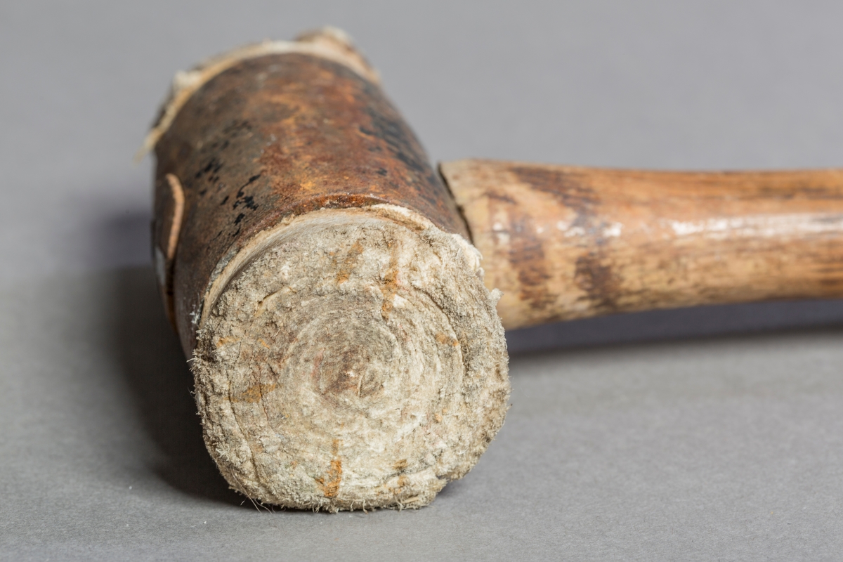 Gjennomgåendetrehåndtak festet med kile og splint til hodet. Hode er en jernsylinder med ruller av råhud satt inn i endene. Rullene har vært surret med lintråd utenfor jernet. Surringen er løs på den ene siden og borte på den andre.