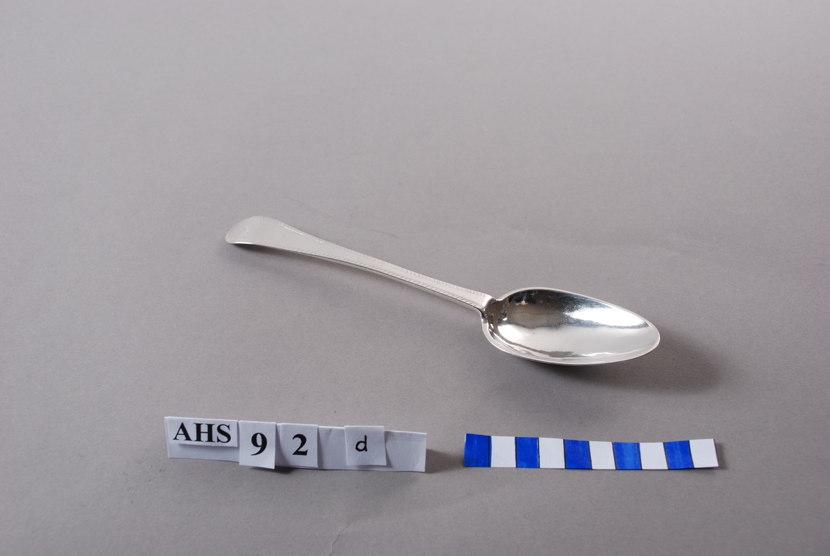 Spiseskje i sølv med produksjonsstempel og guardeinstempel. Skaftet dekorert med en inngravert fjærbord samt bokstaven "K" øverst.