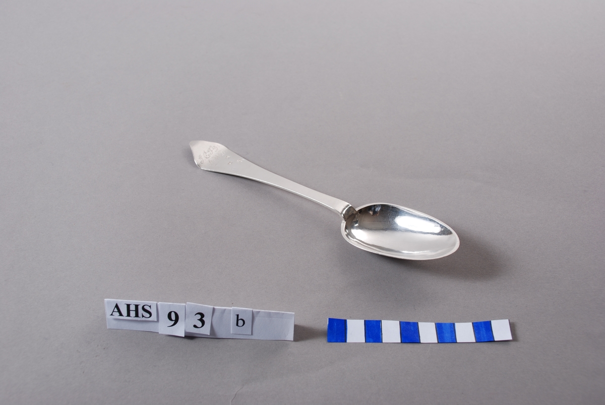 Spiseskje i sølv med stempel og inngraverte initialer. På baksiden er såvel blad som skaft meget rikt dekorert. Bladet har inngravert senrokokkobladverk, skaftet mer klassisistisk ornamentikk.