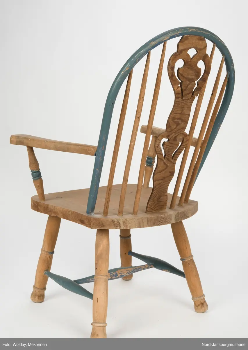 "Budal"inspirert stol, med ryggstø og armelner. Ryggstø er formet av en blåmalt buet list som holdes oppe av seks vertikale trespiler. Den midter, og syvende, spilen er en bred, utskjært list med gjennombrutt mønster, stiliserte hjertemønster. Armlenene er buet utover, og ben og detaljer har dreid dekor.