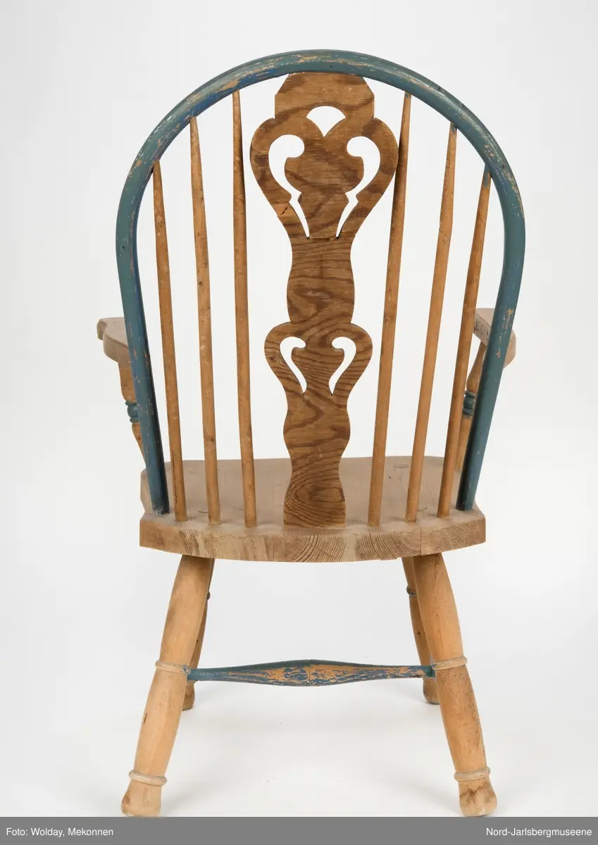 "Budal"inspirert stol, med ryggstø og armelner. Ryggstø er formet av en blåmalt buet list som holdes oppe av seks vertikale trespiler. Den midter, og syvende, spilen er en bred, utskjært list med gjennombrutt mønster, stiliserte hjertemønster. Armlenene er buet utover, og ben og detaljer har dreid dekor.