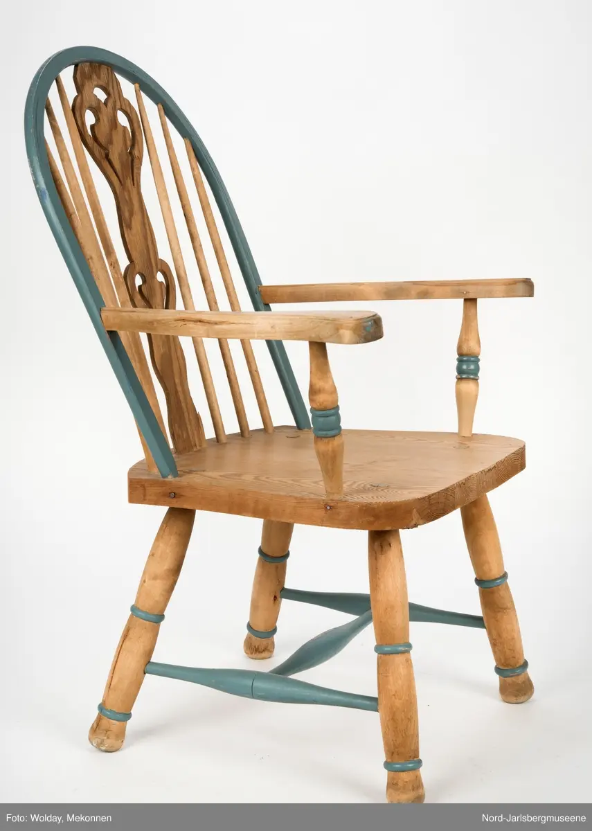 "Budal"inspirert stol, med ryggstø og armelner. Ryggstø er formet av en blåmalt buet list som holdes oppe av seks vertikale trespiler. Den midter, og syvende, spilen er en bred, utskjært list med gjennombrutt mønster, stiliserte hjertemønster. Armlenene er buet utover, og ben og detaljer har dreid dekor.