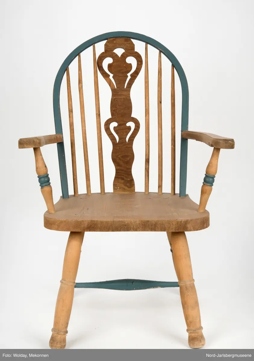 "Budal"inspirert stol, med ryggstø og armelner. Ryggstø er formet av en blåmalt buet list som holdes oppe av seks vertikale trespiler. Den midter, og syvende, spilen er en bred, utskjært list med gjennombrutt mønster, stiliserte hjertemønster. Armlenene er buet utover, og ben og detaljer har dreid dekor.