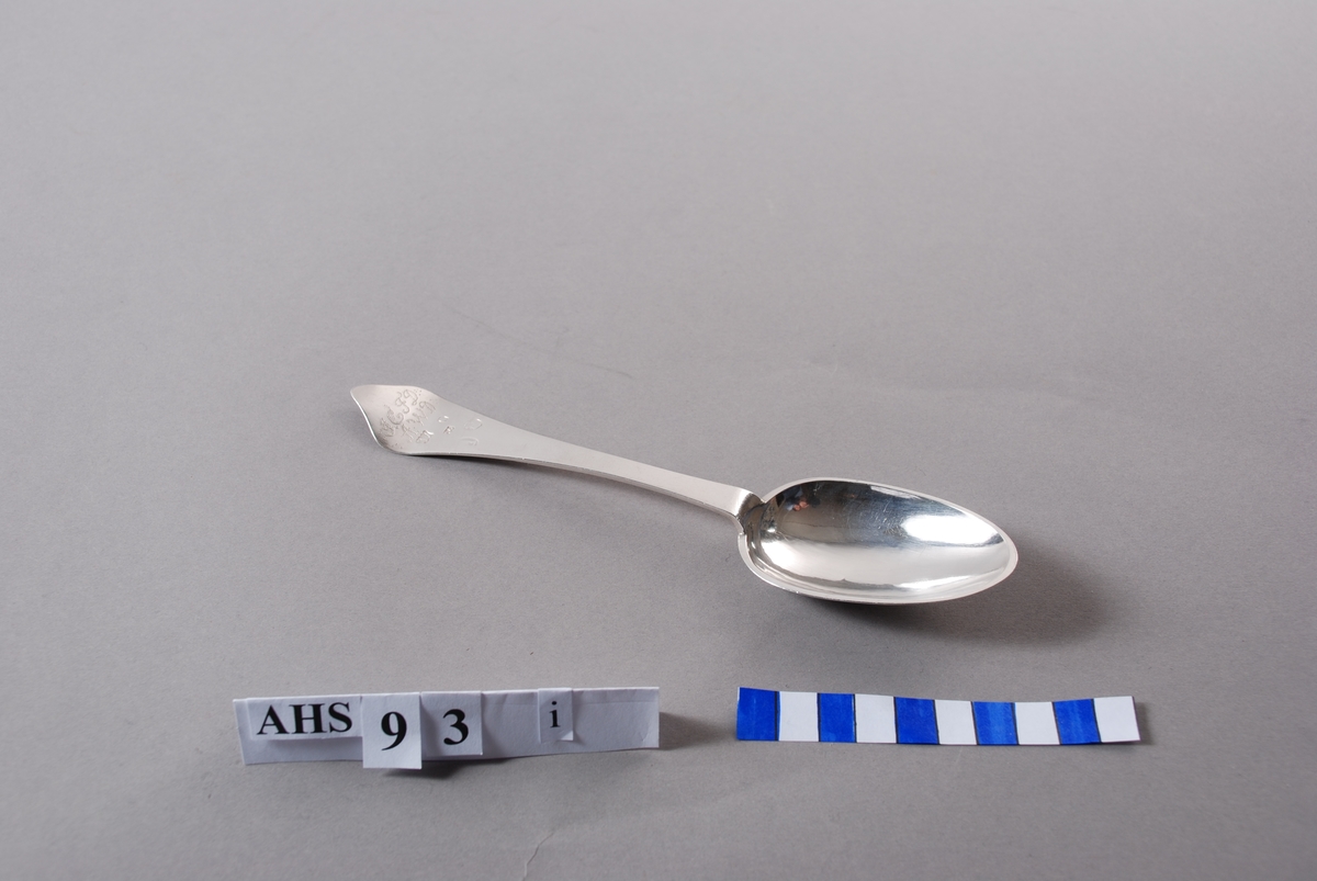 Spiseskje i sølv med stempel og inngraverte initialer. På baksiden er såvel blad som skaft meget rikt dekorert. Bladet har inngravert senrokokkobladverk, skaftet mer klassisistisk ornamentikk.