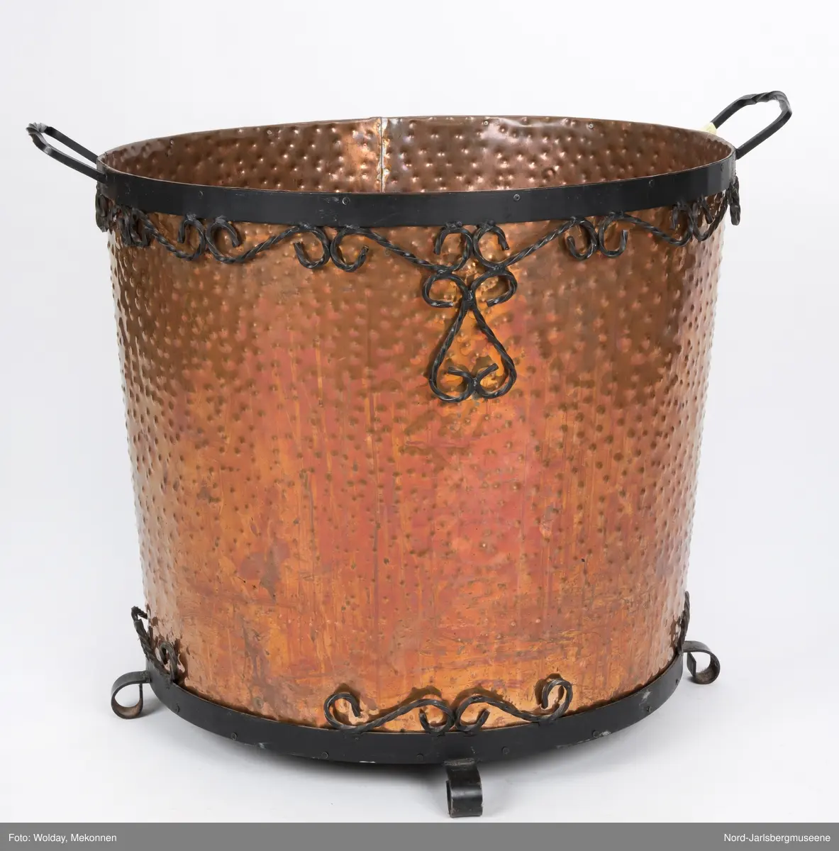 Stor kobberkjele i banket kobber med smijernsdekor i motstilte karnissformer. Smijernsbeslag rundt øvre kant og rundt bunnen, fire små smijernsben. På fronten er festet en messingplate med rød og grønn påskrift