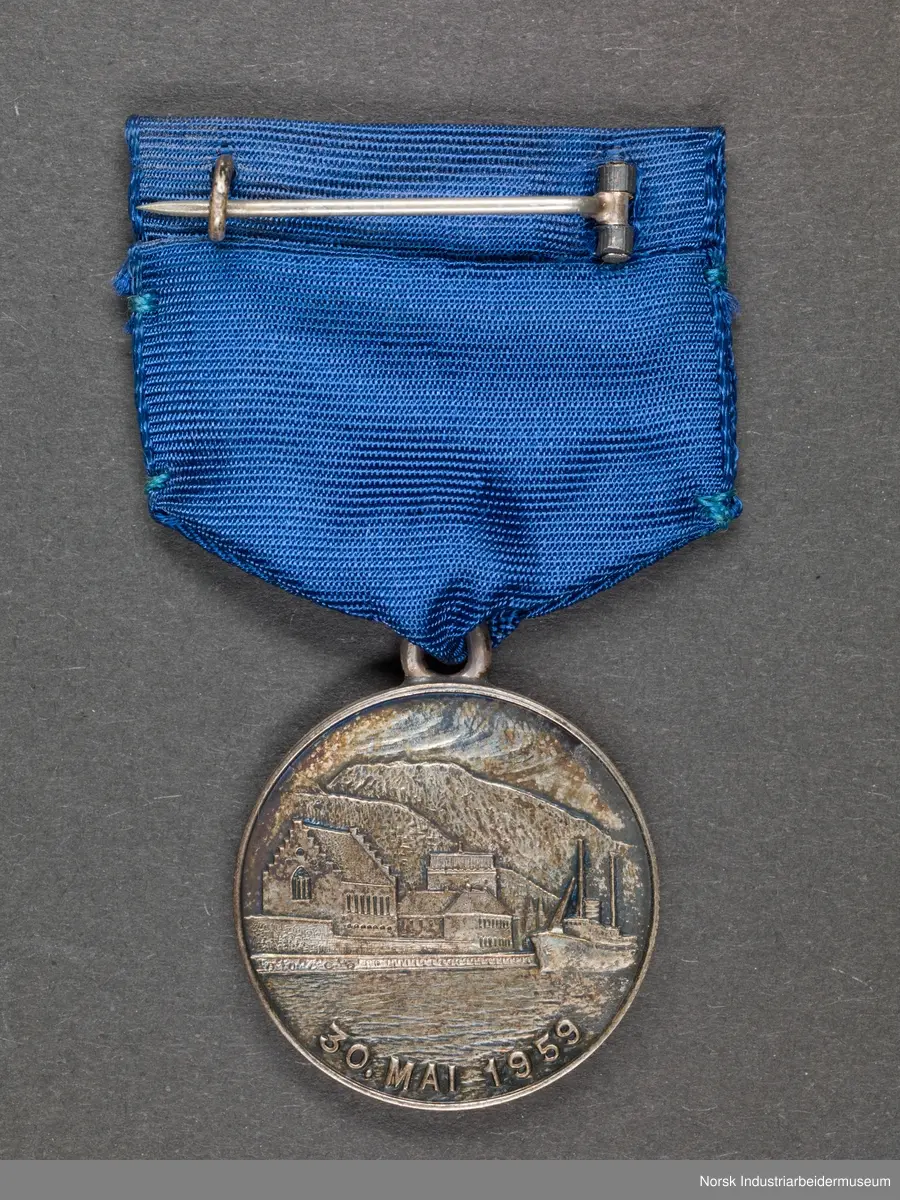 Rund medalje festet til blå tekstil. Nål festet til tekstil. På den ene siden av medaljen er det tekst og emblem med "NGF", tang, ambolt og et kjede. På den andre siden er det motiv av en havn med fjelllandskap i bakgrunnen.