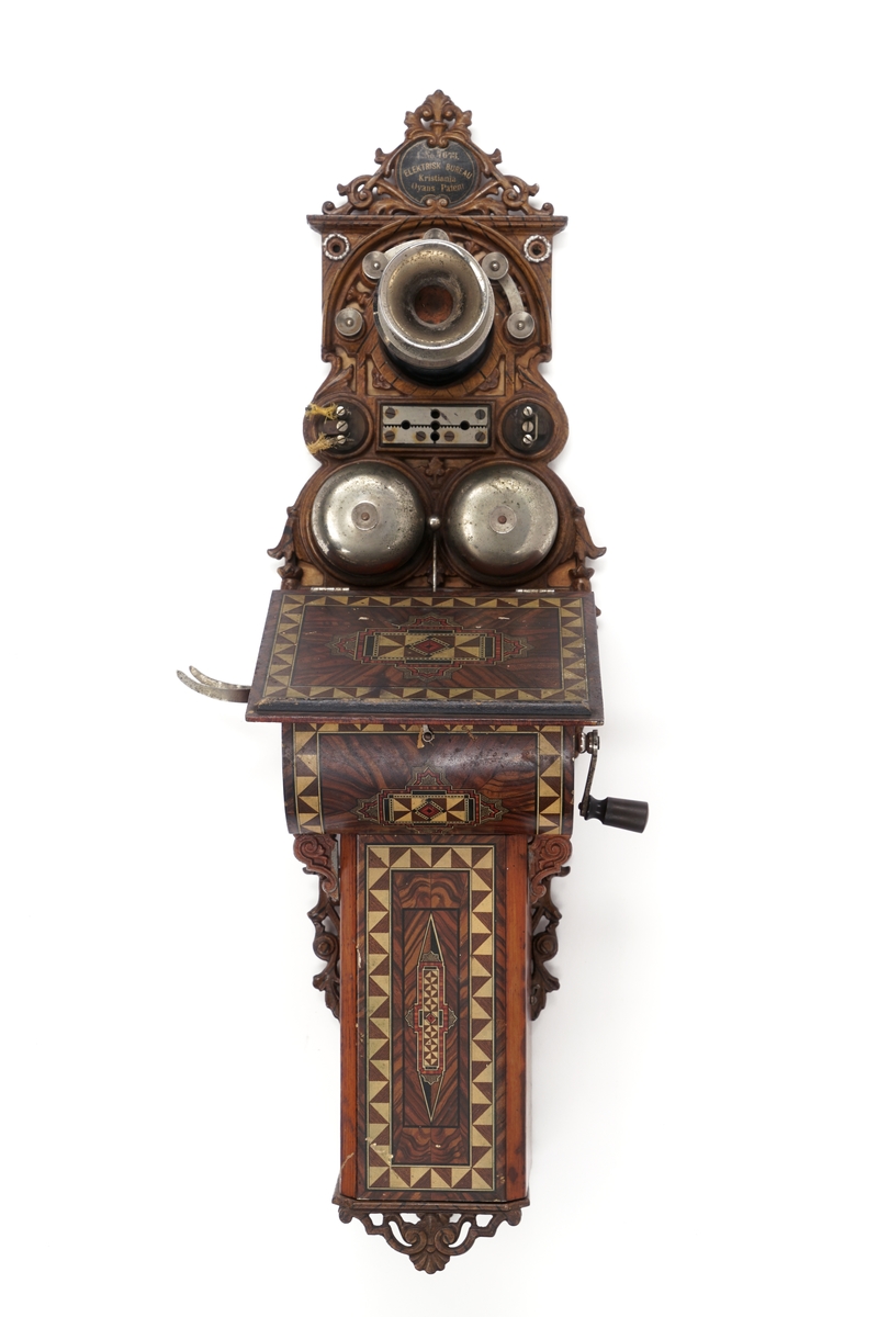 Telefonapparat for kobling på vegg. Fastmontert mikrofon. Høyttaler mangler. Avtagbare, brunlakkerte metalldeksler med dekor. Hengslet topplokk. Smijernsdetaljer. Sveiv med dreid trehåndtak. To bjeller av metall med slagkule mellom disse. Når man dreier på sveiven vil slagkulen "slå" på bjellene og lage en "ringelyd". Diverse koblingspunkter/skruer for tilkobling av ledning.