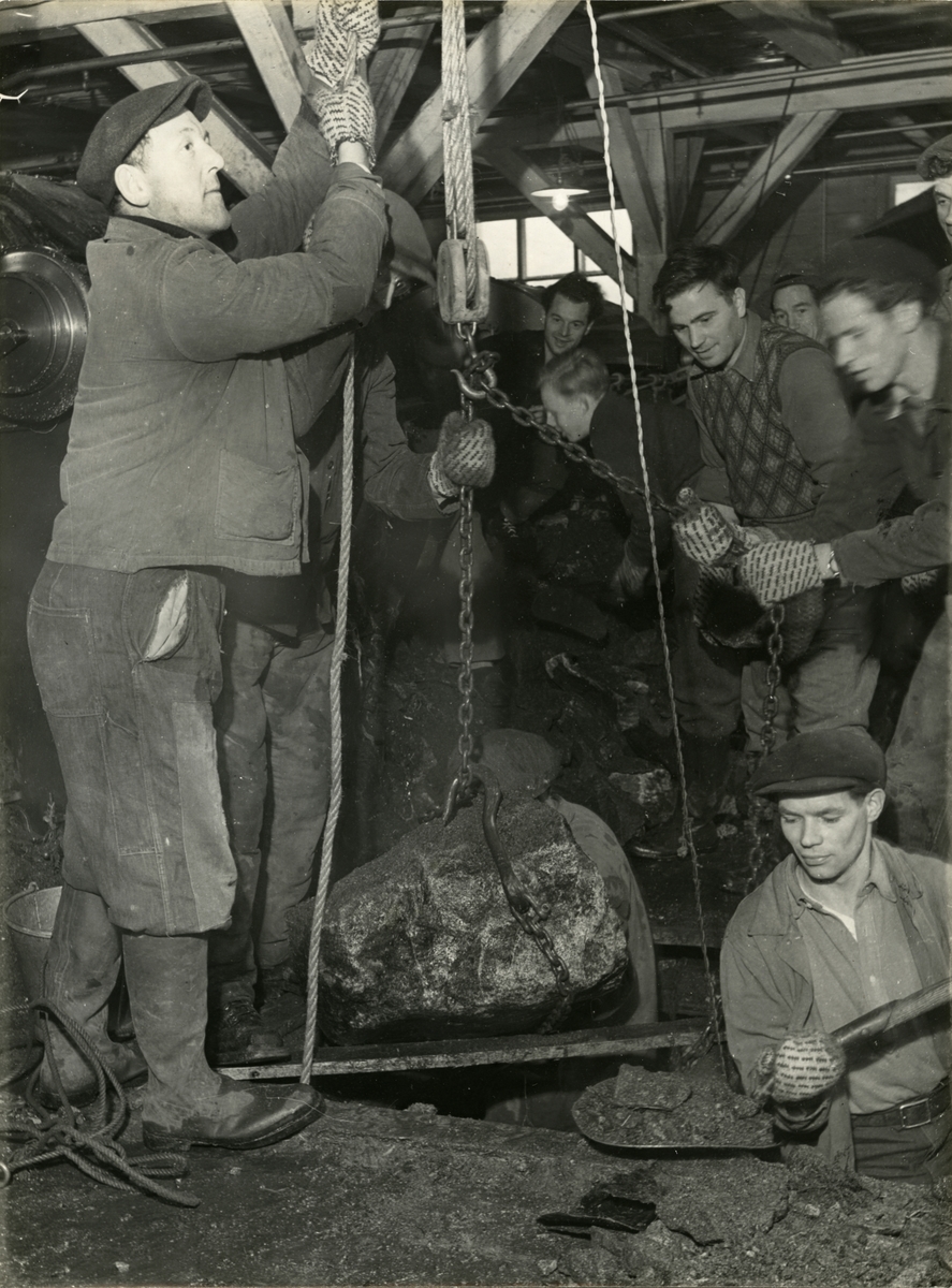 Oppheising av stein etter flaum i Salhus Tricotagefabrik i 1948. Olav Falkanger, murar ved fabrikkanlegget, fremst til venstre i biletet. Arbeidsvottane fleire på biletet går med er laga i avskjer frå islenderstoff.