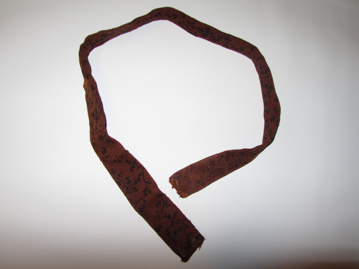 Belte i ull, trykt med svart mønster.  Dobbelt med saum på baksida.  To hekter i kvar ende.  Handsydd. Samansydd av tre delar.