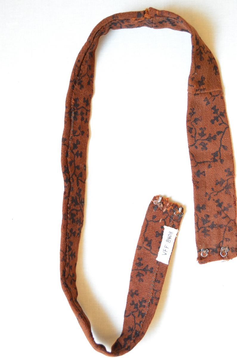 Belte i ull, trykt med svart mønster.  Dobbelt med saum på baksida.  To hekter i kvar ende.  Handsydd. Samansydd av tre delar.