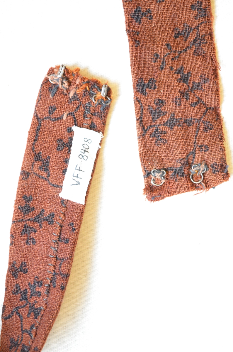 Belte i ull, trykt med svart mønster.  Dobbelt med saum på baksida.  To hekter i kvar ende.  Handsydd. Samansydd av tre delar.