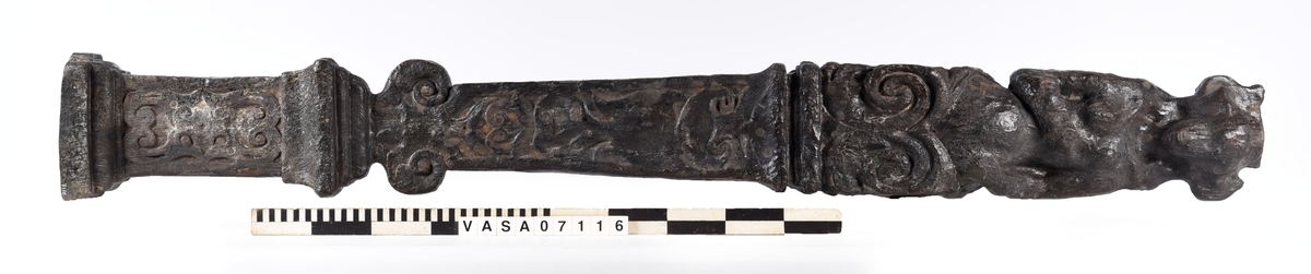 Skulpterad pilaster med övre delen i form av en karyatid med yppig, konstrik frisyr.
Höger arm hålls böjd med handen placerad framför bröstet. Vänster arm hålls utsträckt utefter sidan med handen vilande mot höften. Underkroppen döljs av ett hölje som framtill är prytt med stora, spiralformade ornament och små djurfigurer.
Karyatiden avgränsas från det rullverksornerade pilasterskaftet av en profilerad list. Nedtill avslutas skaftet av ett rullverksornament ovanför den rikt profilerade basen. Nedtill är pilastern försedd med en hög piedestal, prydd med beslagsornamentik. Figuren och dekoren omfattar även pilasterns sidor. Baksidan är slät.


Text in English: Sculpted pilaster with its upper part shaped as a Caryatid with a luxuriant artistic coiffure.
The right arm is held bent with the hand in front of the breast, the left arm is held at the side with the hand resting against the thigh. The lower part of the body is concealed by a shift or slip that is decorated with large spiral-shaped ornaments and small figures of animals.
A profiled batten separates the Caryatid from the shaft of the pilaster, which is ornamented with scrollwork. The shaft terminates with a scrollwork ornament above the richly profiled base. Below, the pilster is equipped with a high pedestal, decked with applied ornamentation. The back is smooth.