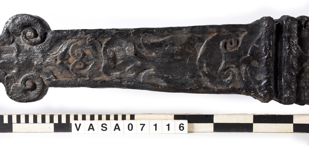 Skulpterad pilaster med övre delen i form av en karyatid med yppig, konstrik frisyr.
Höger arm hålls böjd med handen placerad framför bröstet. Vänster arm hålls utsträckt utefter sidan med handen vilande mot höften. Underkroppen döljs av ett hölje som framtill är prytt med stora, spiralformade ornament och små djurfigurer.
Karyatiden avgränsas från det rullverksornerade pilasterskaftet av en profilerad list. Nedtill avslutas skaftet av ett rullverksornament ovanför den rikt profilerade basen. Nedtill är pilastern försedd med en hög piedestal, prydd med beslagsornamentik. Figuren och dekoren omfattar även pilasterns sidor. Baksidan är slät.


Text in English: Sculpted pilaster with its upper part shaped as a Caryatid with a luxuriant artistic coiffure.
The right arm is held bent with the hand in front of the breast, the left arm is held at the side with the hand resting against the thigh. The lower part of the body is concealed by a shift or slip that is decorated with large spiral-shaped ornaments and small figures of animals.
A profiled batten separates the Caryatid from the shaft of the pilaster, which is ornamented with scrollwork. The shaft terminates with a scrollwork ornament above the richly profiled base. Below, the pilster is equipped with a high pedestal, decked with applied ornamentation. The back is smooth.