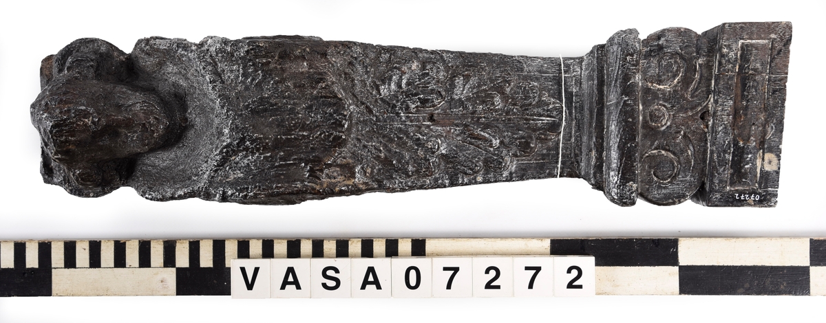 Liten skulpterad pilaster, utformad som en karyatid eller möjligen en putto. Figuren har långt hår och ett knubbigt ansikte, omgivet av en bred krage. Det nedåt avsmalnande pilasterskaftet är dekorerat med akantusblad. Nedtill avslutas pilastern av en rikt profilerad och dekorerad sockel. På skulpturens släta baksida sitter en nygjord upphängningsanordning, fästad med två skruvar.
Skulpturen är sliten.

Text in English: A small carved pilaster, with a caryatid or probably a putto. The figure has a chubby face, long hair and a broad collar. The lower part of the pilaster is decorated with acanthus leaves. The caryatid stands on a richly profiled and decorated socle. On the smooth back of the sculpture is a new hanging device attached with two screws.
The sculpture is worn.