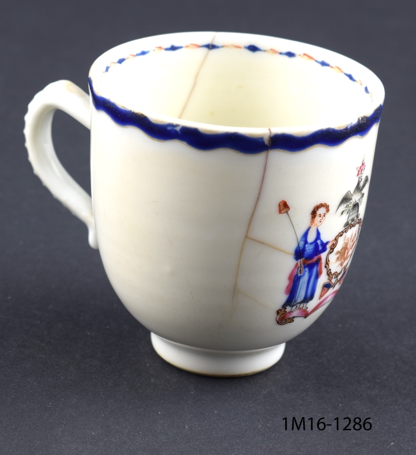 Vit kopp med blå kanter upptill. Motiv mitt på koppen med två kvinnor och en fågel.