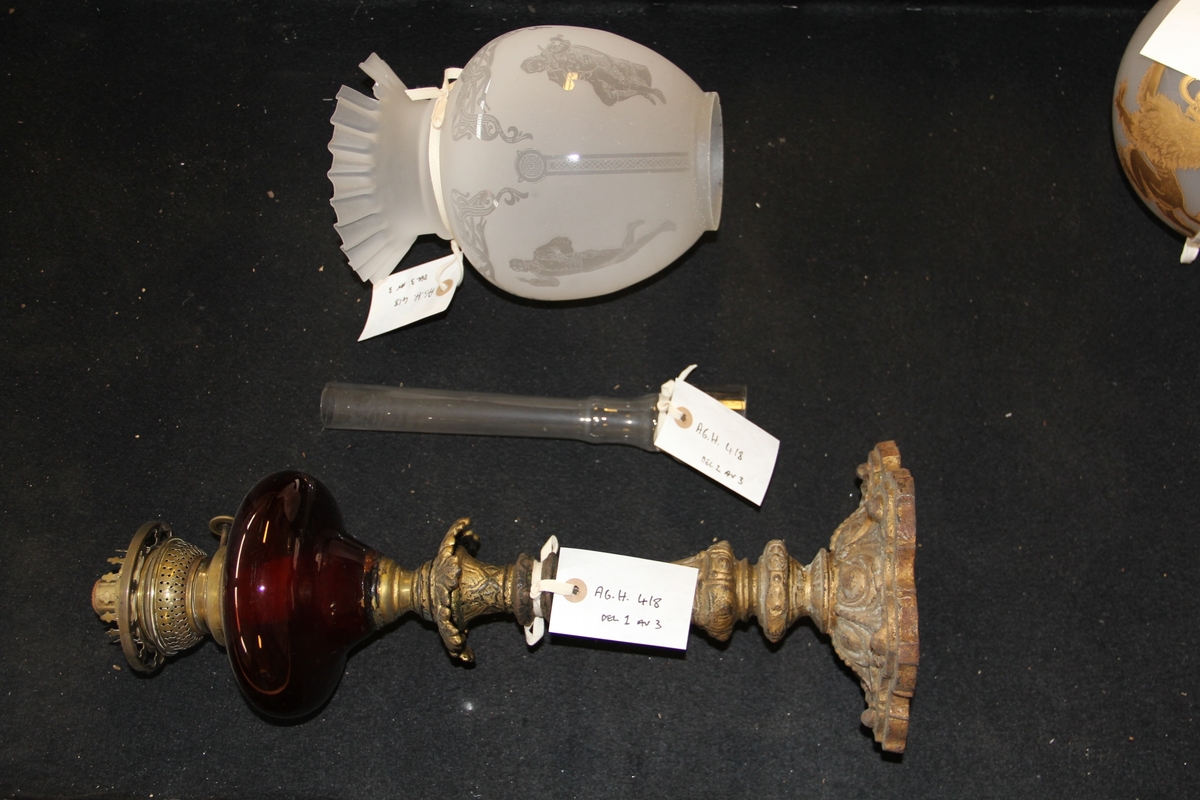 Parafinlampe, foten er støpt av messing med en mørkerød beholder og hvit kuppel av glass. Kuppelen er dekorert med dragemotiv, spillemann og bunadskledde figurer.