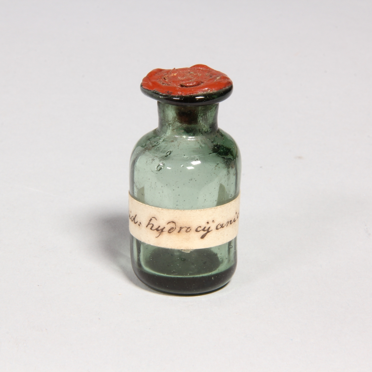 Flaskor, 10 st, av grönt glas med proppar av kork förseglade med rött lack. Står i pappask med lock klädd med marmorerat papper. Flaskorna innehåller rester av cyanid.