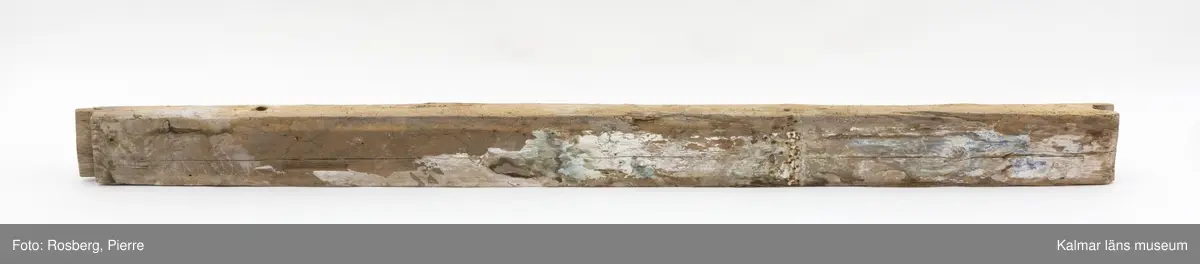 KLM 17275:4:1. Vägg, del av. En timmerstock tillhörande en innervägg. Hål för trädymlingar. På båda sidor bevarade fragment av bemålning, troligen från 1600-talet. På ena sidan måleri i bl.a. vitt och blått. På andra sidan bemålning i vitt, blått och grönt samt rester av påspikade tapeter. Även text skrivet med blyerts, något otydlig men troligen: Lundgren.