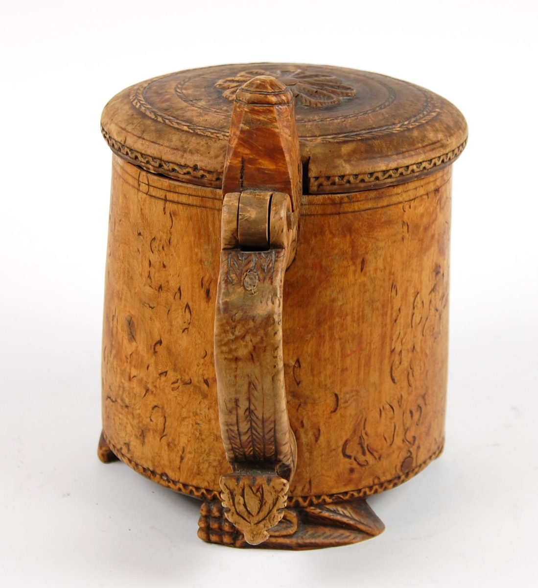 Kanne av dreid valbjørk. Lokk med skjært rosett og to bårder. Tre føtter med utskjæringer.