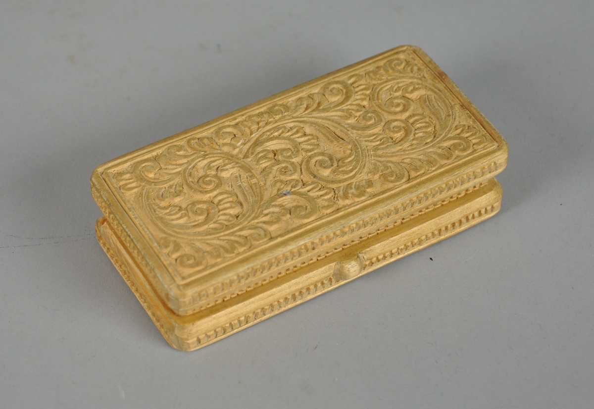 Liten eske av tre, med skjært dekor. Esken har en rektangulær utforming og skjært dekor på lokk og bunn med akantusmotiv, og linjer/rutenett på sidene. 

Esken har en knott på framsiden av lokket.