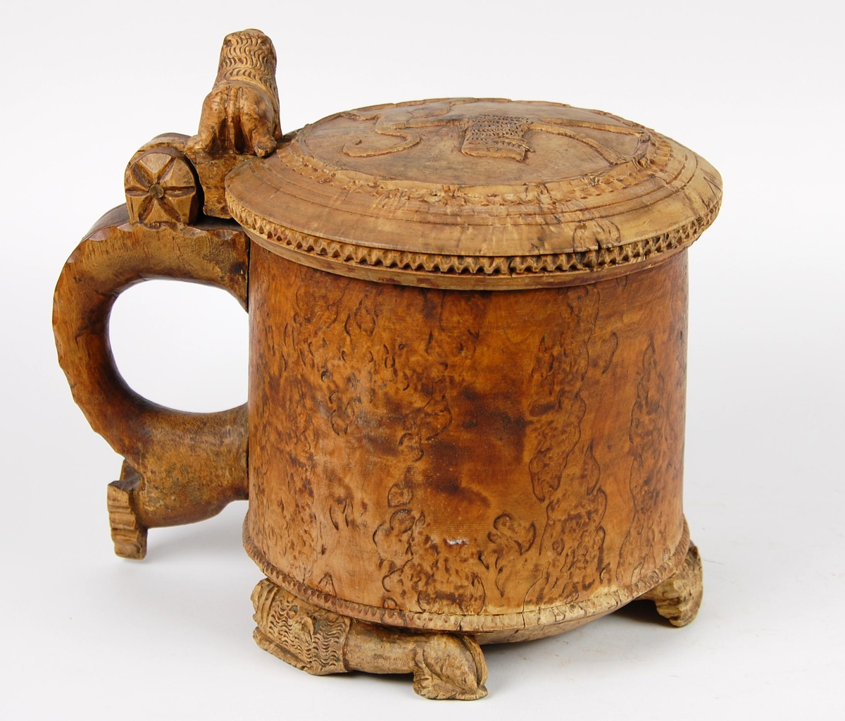 Kanne av dreid valbjørk. Lokket med uskåret springende løve omgitt av bård, bølgete utskjæringer langs kanten. Lokkåpner formet som en løve. Hanken med skjæringer langs kanten, ender i en spiss. Tre liggende løver danner føttene. Buemerke under.