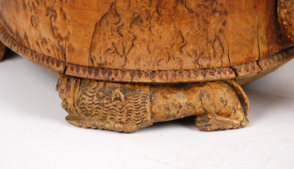 Kanne av dreid valbjørk. Lokket med uskåret springende løve omgitt av bård, bølgete utskjæringer langs kanten. Lokkåpner formet som en løve. Hanken med skjæringer langs kanten, ender i en spiss. Tre liggende løver danner føttene. Buemerke under.