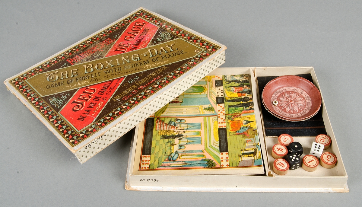 Sällskapsspel "The boxing-day. Game of forfeit with redeem of pledge. The 25 th December". I kartong med tryck i rött, guld och brunt. Till spelet hör: 1 spelplan av vaxat papper, färgtryck på väv, 6 runda spelbrickor, 3 tärningar, 2 svarta av trä, 1 vit av porslin, 2 små pappersskålar, 1 liten grå krita och 1 griffeltavla av papp, samt en spelbeskrivning.