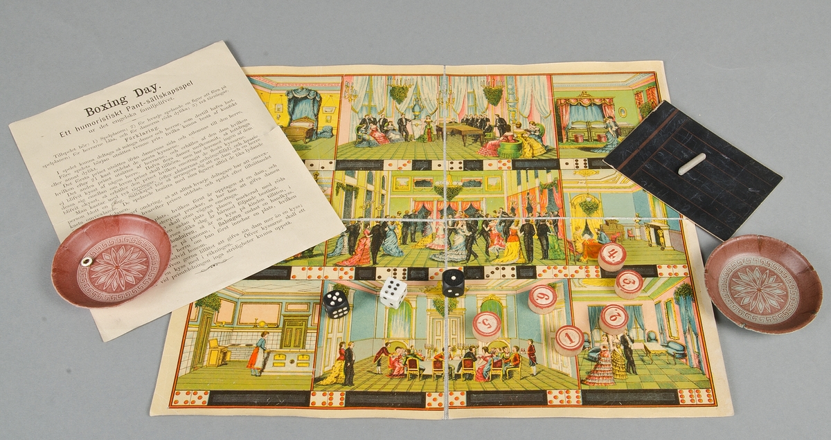 Sällskapsspel "The boxing-day. Game of forfeit with redeem of pledge. The 25 th December". I kartong med tryck i rött, guld och brunt. Till spelet hör: 1 spelplan av vaxat papper, färgtryck på väv, 6 runda spelbrickor, 3 tärningar, 2 svarta av trä, 1 vit av porslin, 2 små pappersskålar, 1 liten grå krita och 1 griffeltavla av papp, samt en spelbeskrivning.