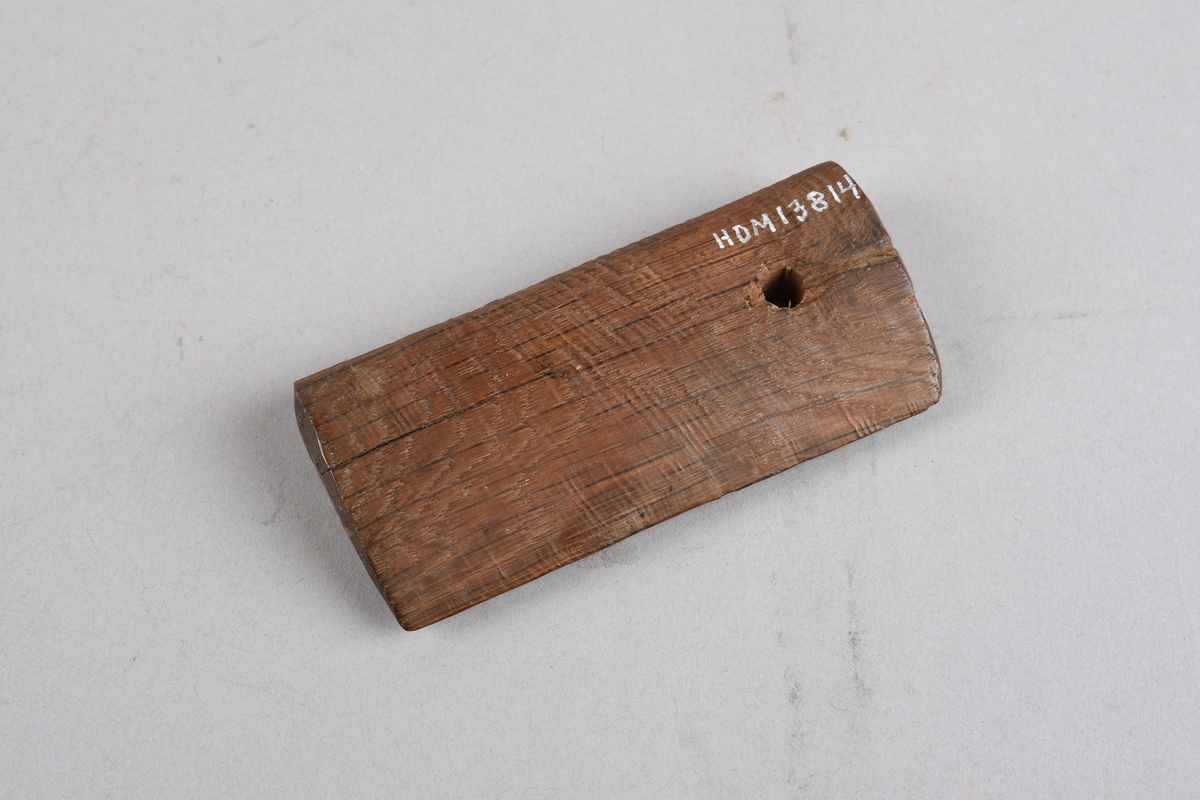 Avlangt trestykke med dråpeforma tverrsnitt, skarp kant på eine langsida og runda på andre.Hol gjennom trestykket, spor i treet frå holet ut til kortenden. Laga i mørkt treverk, ubehandla.