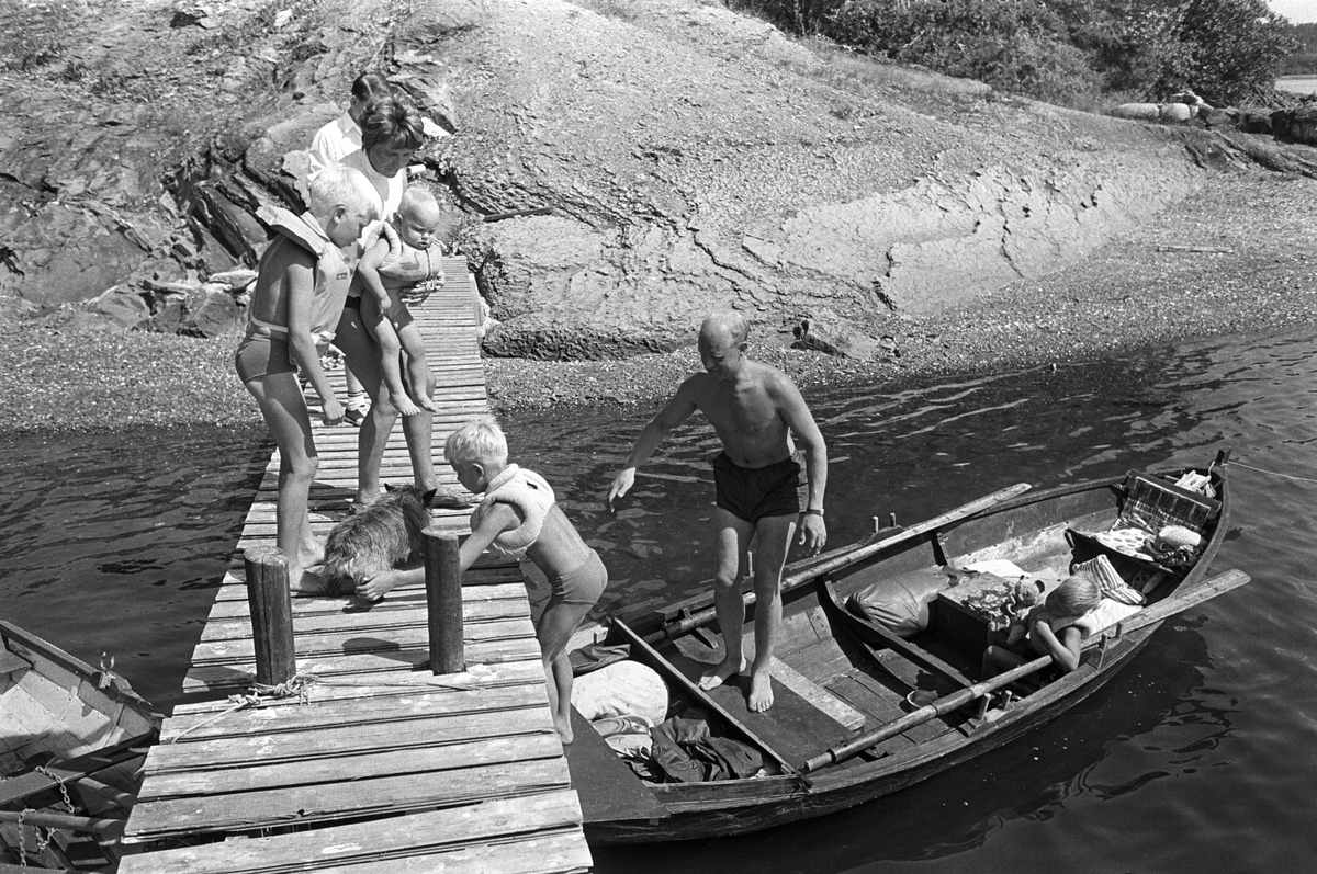 Erik Damman, reklamekonsulent fra Oslo, ferierer i båt med familien i Oslofjorden. De skal ro 10 - 12 mil i løpet av 8 - 10 dager, og går i land på små holmer som fjorden har mange av. Like før avreise fra Brønnøya. Ragnhild Damman på brygga med Jaran (1,5) og Rein (10). Are (8) med hunden Rocky. I båten Erik Damman og Brede (5).