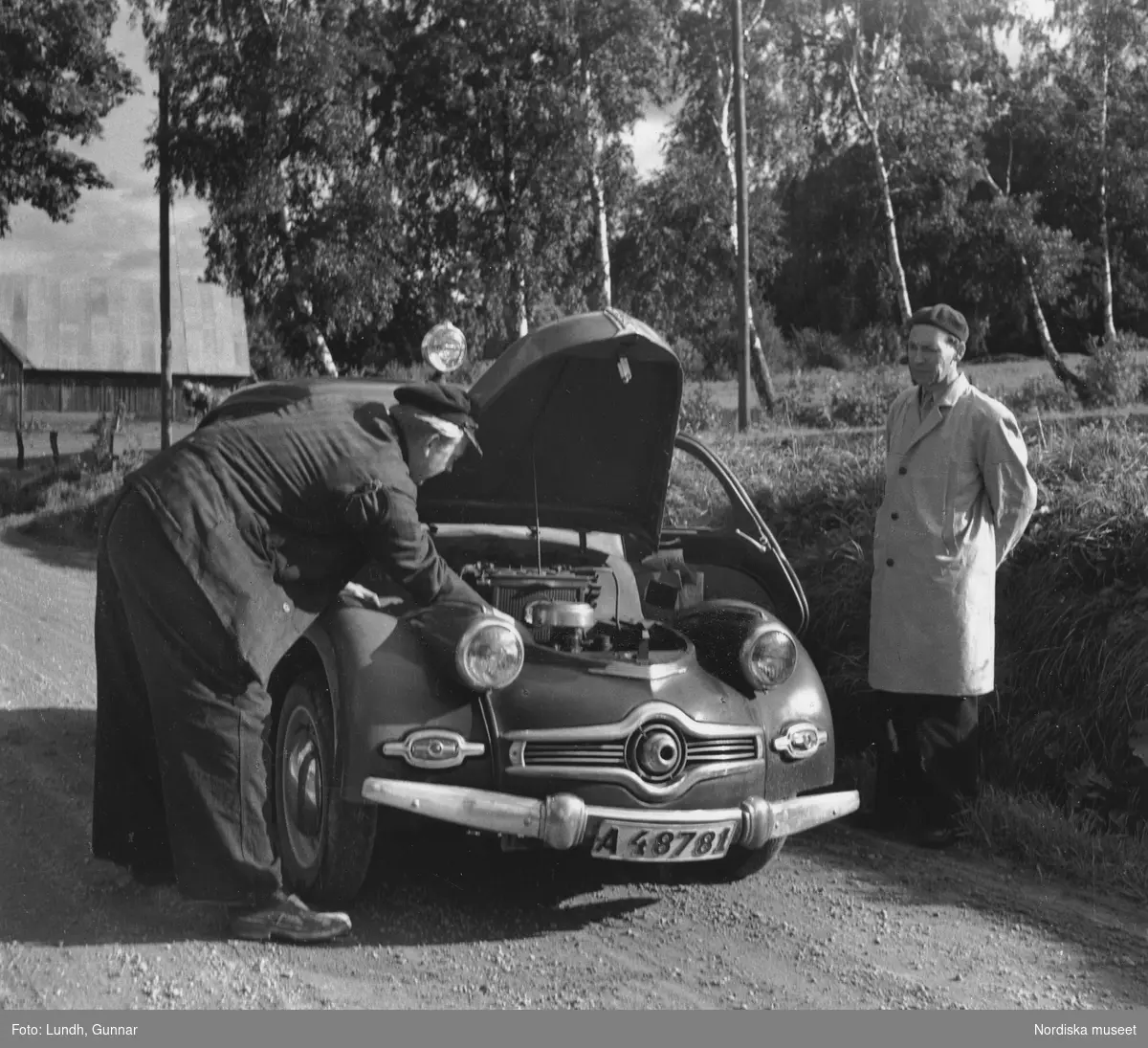 Fotografen Gunnar Lundhs bil, en Dyna Panhard med uppfälld motorhuv på grusväg. En man böjd över motorn, en annan står intill och betraktar arbetet.