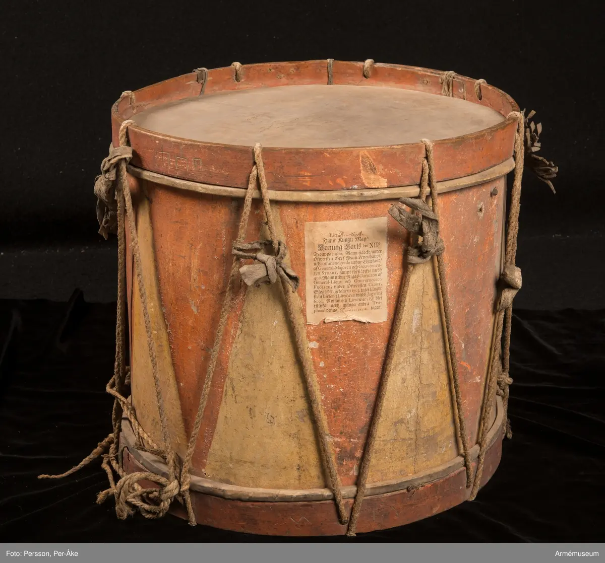 Cylindertrumma av trä med bemålad lim och krita grunderad duk.
Ytan är krackelerad med ett visst bortfall, dock relativt stabil. Tre hål i varierande storlek.

Membran: övre skinnet är smutsigt och repigt med tendenser till sprickbildning. Det finns en centimeterstor lagning. Det undre, har en tvärsgående spricka som tudelar skinnet. Den större delen rullar in mot mitten. Det finns fläckar av ngn. lättlöslig, gråaktig lösning. 

Veckelgjordar: har lindade fogar, den övre med tunt rep/lina, 
Den undre med rep och smalt läderband. Tre tecken inskurna i övre veckelgjordens utsida, som liknar: 1:a,  gaffel med tre klor, 2:a, endast de två yttre klorna och skaft, 3:e tecknet: endast de två yttre klorna, skaftet saknas. 

Spännlina: är fin med tvinnat, avsmalnande repslut. Det finns åtta spännstroppar varav två stycken är fransade.

Spännringar : Utsmyckning : röda kilar målade på gul botten. Mönstret ristat i grunden.

Märkning: fastklistrad Konung Karl den XII med ifyllnad: Litt B  No.4 
Hängandes pappers etikett  i påse No 1211,
Gustavianska koret No 9.