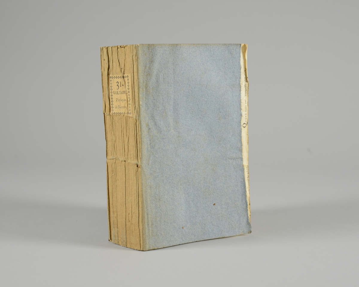 Bok, pappband,"Oeuvres completes de Voltaire", del 31 "Philosophie de Newton", tryckt 1785.
Pärmen klädd med gråblått papper, på pärmarnas insidor klistrade sior ur annan bok. Med skurna snitt. På ryggen klistrad pappersetikett med tryckt text samt volymens nummer. Ryggen blekt. Med planscher.
