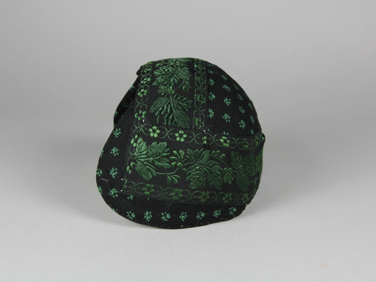 Bindmössa till Häverödräkten. Stomme av vit stärkt tuskaftsvävt tyg som sytts ihop av tre delar och pressats. Klätt med svart bomullstyg med tryckt grönt mönster av små blommor. Runt hela mössan är ett 5 cm brett sidenband fastnålat. Bandet är svart med grönt mönster. Mössans kanter är stickade med prickstygn i två rader.