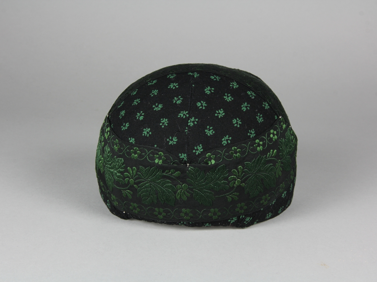 Bindmössa till Häverödräkten. Stomme av vit stärkt tuskaftsvävt tyg som sytts ihop av tre delar och pressats. Klätt med svart bomullstyg med tryckt grönt mönster av små blommor. Runt hela mössan är ett 5 cm brett sidenband fastnålat. Bandet är svart med grönt mönster. Mössans kanter är stickade med prickstygn i två rader.