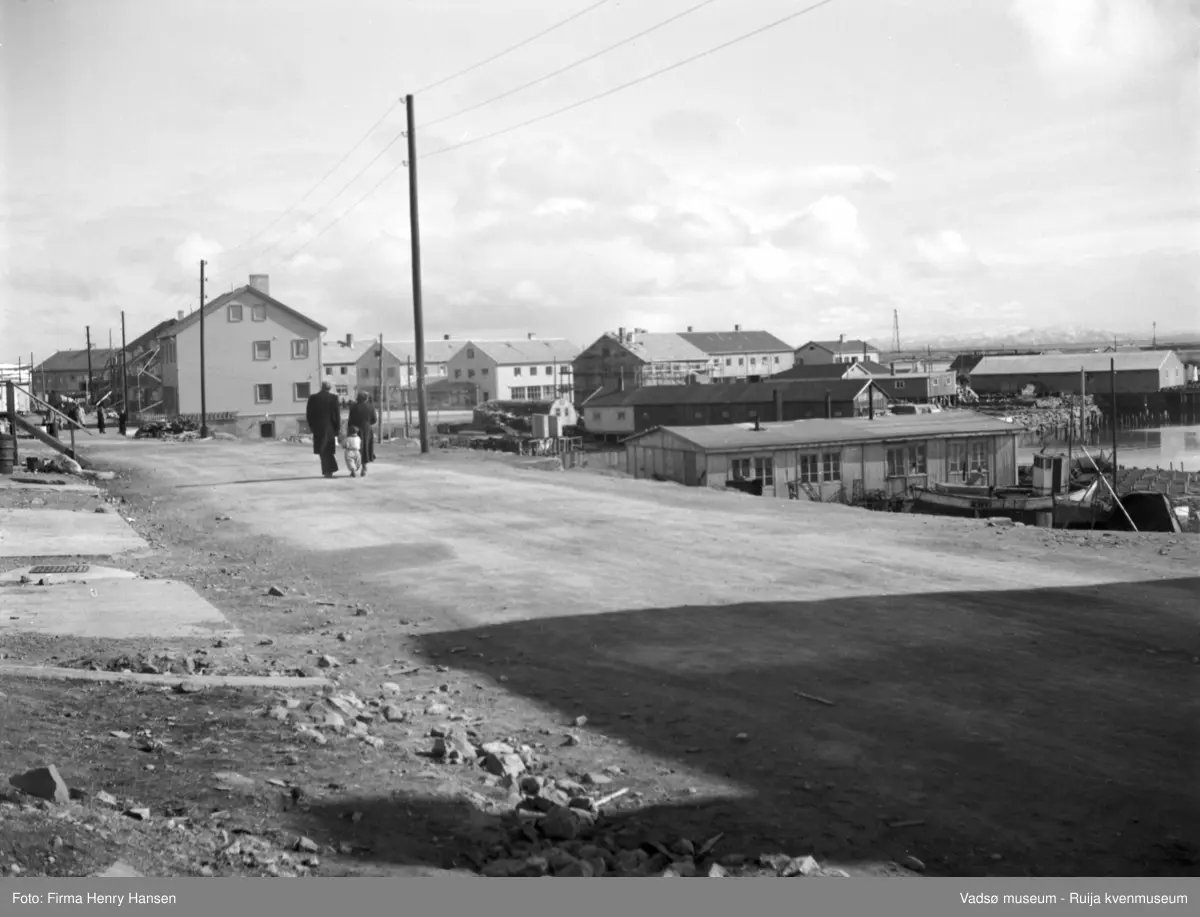 Vadsø mai 1951. Havnegata østover, med gjenreisningshus. Flere av byggene rundt Torget og langs Tollbugata er ferdige, stillas fremdeles rundt ett av byggene.  Nærmest fotografen er ei brakke, flere brakker i bakgrunnen. Man ser også Vadsøya  med luftskipsmasta.