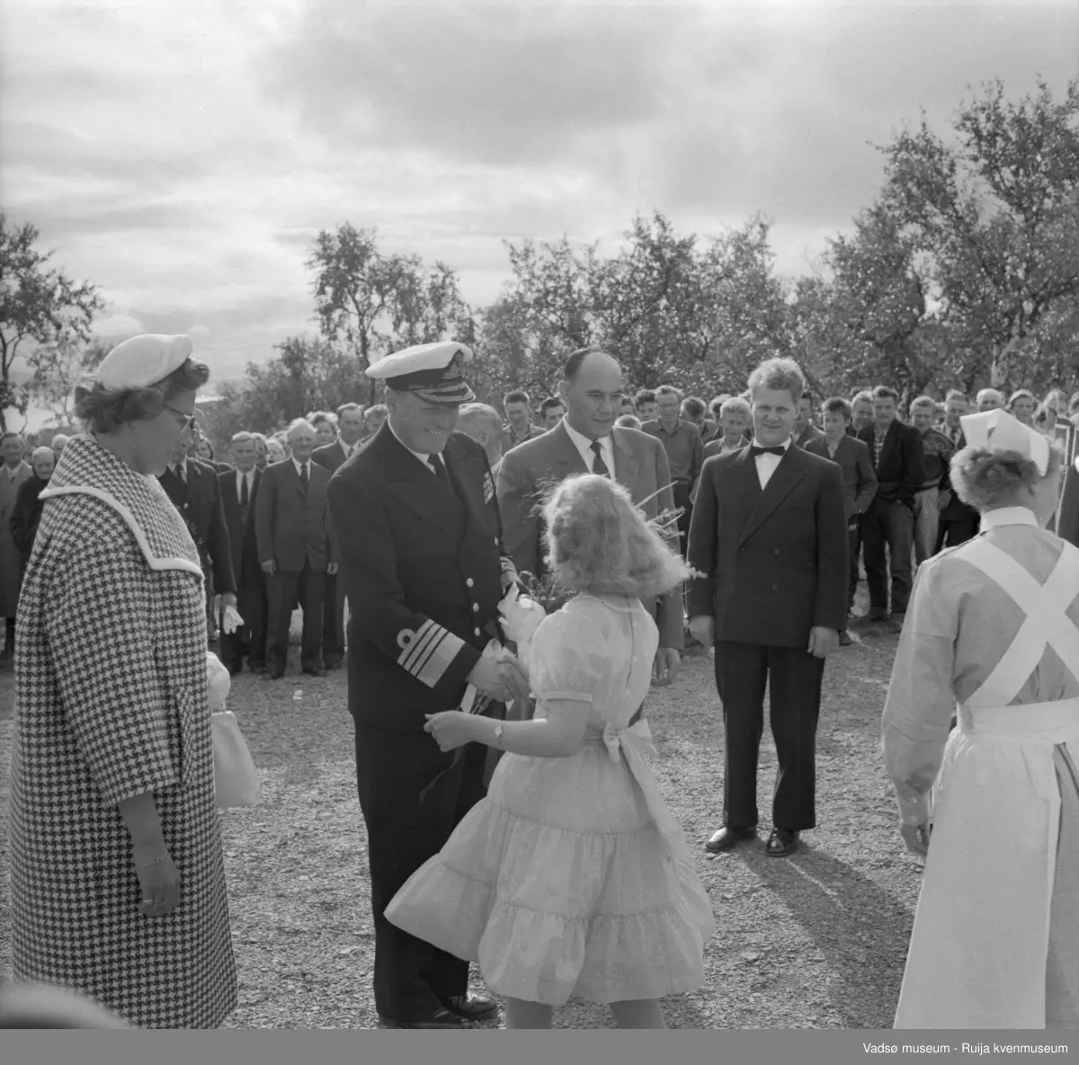 Vestre Jakobselv juli 1959. Kongebesøk på barnehjemmet Vårsol. Kongen får overrakt blomster av Anne Marie Sommer. Til venstre for Kongen prinsesse Astrid. Til høyre for Kongen ordfører Henry Karlsen, Anaton Strand og søster Gunvor. Mange av bygdas barn og voksne i bakgrunnen.