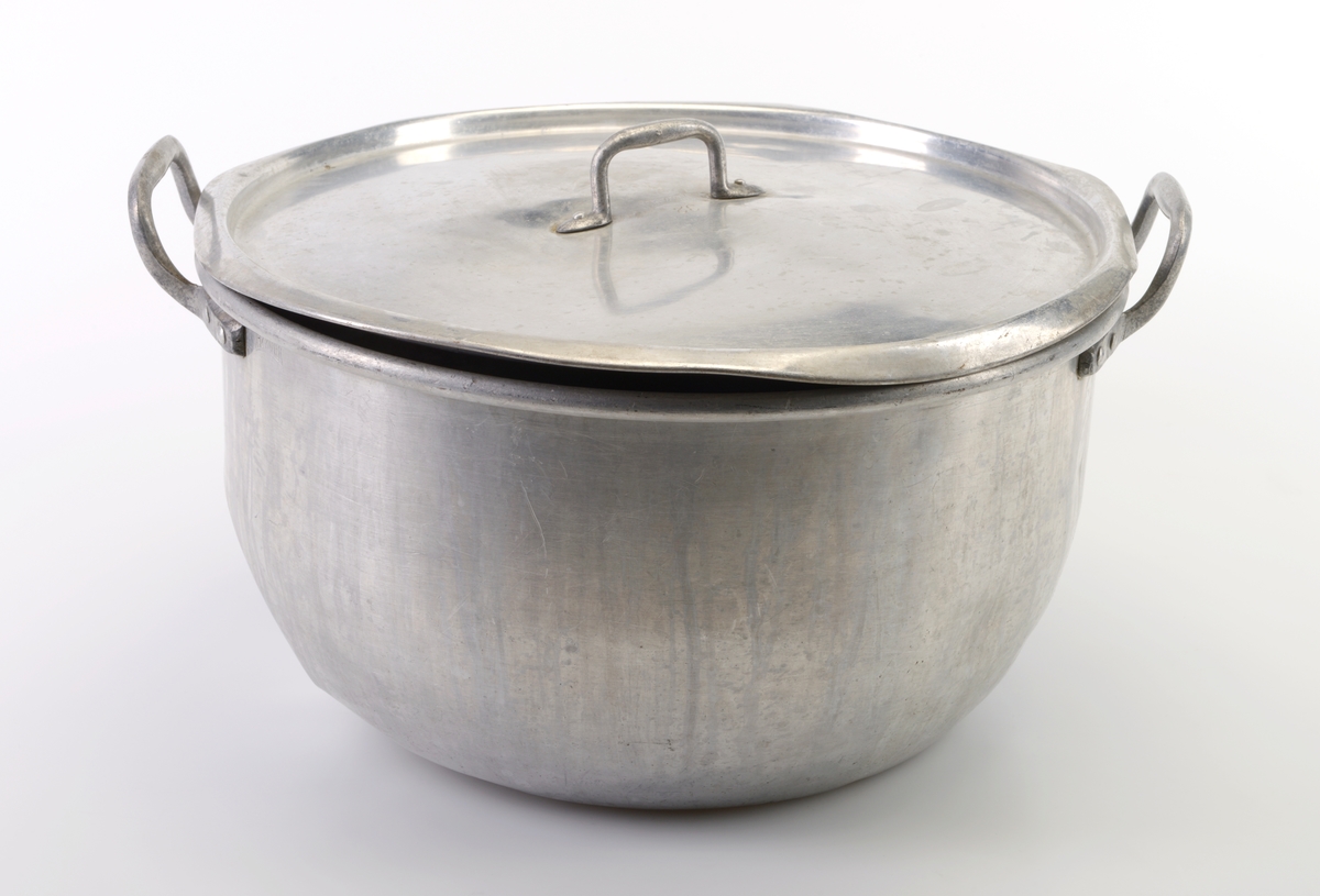 En kasserolle med lokk laget av aluminium. Kasserollen er stor og har buede sider inn mot bunnen. I hver side er det festet en hank (naglet/loddet fast). Det hører til et lokk som også er laget av aluminium. På misten av lokket er det festet et håndtak (naglet/loddet fast).
