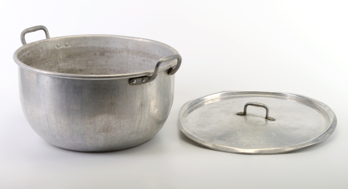 En kasserolle med lokk laget av aluminium. Kasserollen er stor og har buede sider inn mot bunnen. I hver side er det festet en hank (naglet/loddet fast). Det hører til et lokk som også er laget av aluminium. På misten av lokket er det festet et håndtak (naglet/loddet fast).