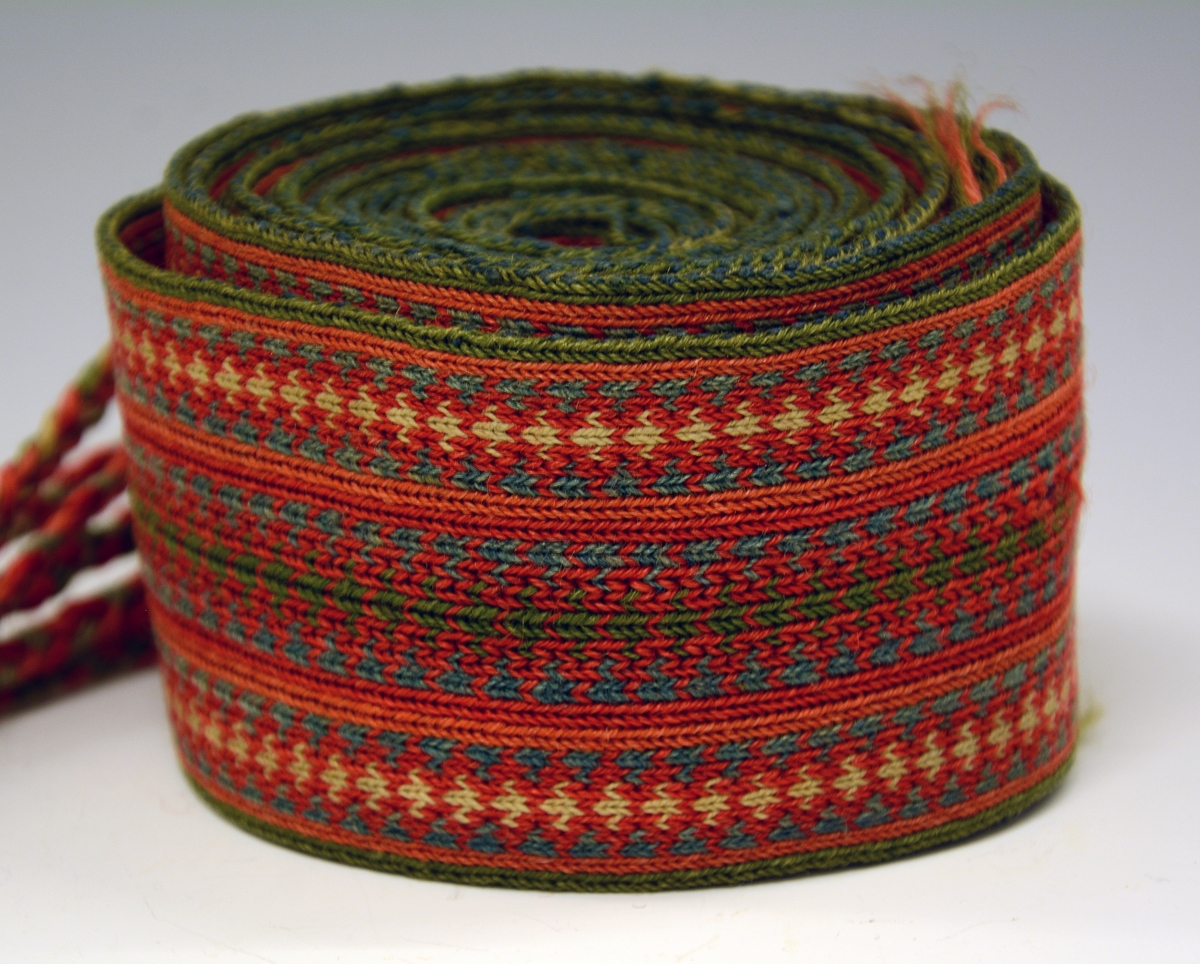 Belte til folkedrakt, brikkevevet. Frynser i den ene enden, de er flettet. Den andre enden er det renning uten avslutning.
