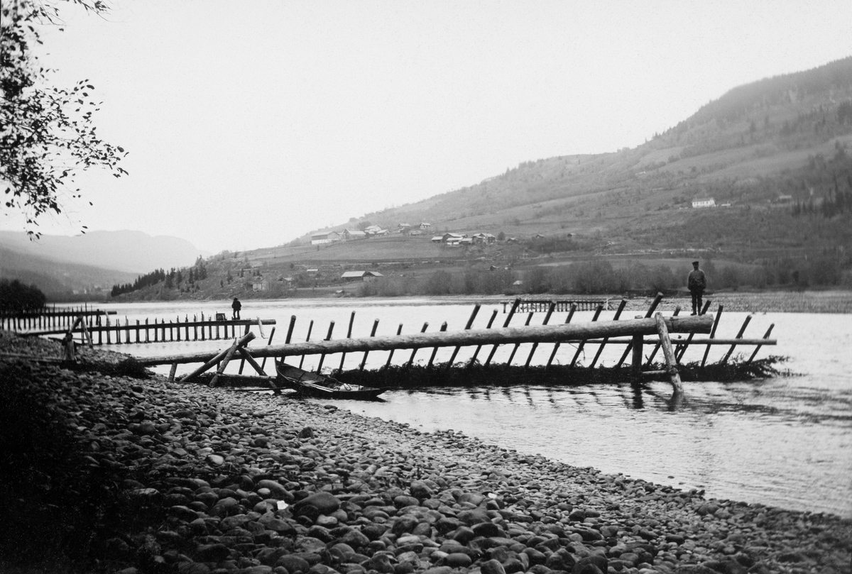 Såkalt «utgjerdstre» ved Knuvelen i nerdre del av Gudbrandsdalslågen, i daværende Fåberg kommune.  Fotografiet skal være tatt i 1904.  Utgjerdstreet var en strømbryterinnretning som skulle muliggjøre notkast på lokaliteter der det kunne være mye i fisk som rant så fort at det kunne bli vanskelig å berge nota.  Ved Knuvelen var det to slike strømbrytere, et «utgjerdstre» øverst og et «landingstre» lengre nede.  Begge disse innretningene var lagd av lange trestammer som var lagt med rotenden utover i elveløpet og støttet opp av to bein - «stetter» i ytterendene.  I dette tilfellet var det også en «bjønn», en slags bukk av tre, som skulle stabilisere utgjerdstreet.  Fra den lange stokken ble det stukket rajer skrått ned mot elvebotnen på motstrøms side, og mellom disse ble det flettet kvist fra grantrær, som skulle bremse vannstrømmen.  Nota ble kastet fra en båt, ei «åfløy» (båttypen vi her skimter fortøyd ved siden av bjønnen).  Den ble deretter rodd i ei bue utover i elveløpet, og deretter tibake mot elevbredden like ovenfor det nedenforliggende landingstreet.  Her gjaldt det å ha mye mannskap, som kunne trekke nota og fangsten på land før strømmen dro verdiene med seg (jfr. Hartvig Huitfeldt-Kaas' beskrivelse, som er gjengitt under fanen «Opplysninger»).  Opprinnelig fulgte retten til notfiske på dette stedet gardene Røyne, Borud, Søndre Hov, Børke, Onsum og Nordre Jørstad i Fåberg.  Seinere ble det solgt andelsretter til flere mindre «lotter» i fisket.  Notfisket ved Knuvelvarpet opphørte etter mjøsregukleringa i 1965.  Fra da av ble strømmen i Lågen for svak på dette stedet.  

I bakgrunnen skimter vi «mækrakker».  Dette var utgjerdstrær hvor det var montert tett i tett med ruser som skulle fange fisk som var på vandring oppover elveløpet (jfr. f. eks. SJF.1988-00312 og SJF.1988-00313).