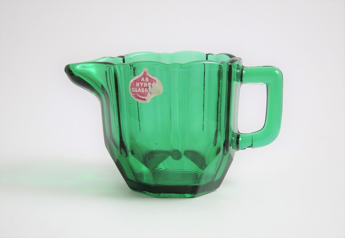 Gräddsnipa med pip och grep.
Runt om facett- och skärslipimitation. 
Etikett: Lila botten med guldfärgad ram och text: AB NYBRO GLASBRUK.