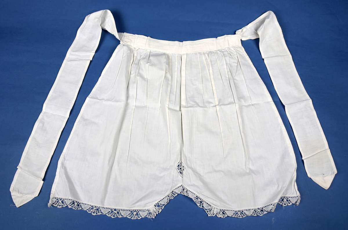 Forkle med påsydd blondekant nederst. Nedkanten på forkleet går opp i en spiss på midten.