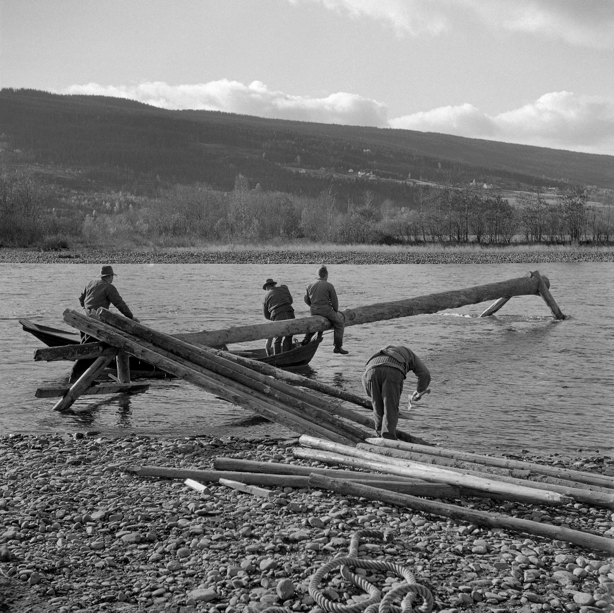 Dette fotografiet viser klargjøring av en strømbrytende innretning – et utgjerds- eller landingstre – i Gudbrandsdalslågen ved Øyra i Fåberg i 1962. Slike innretninger ble lagd for å muliggjøre notkast etter lågåsild (Caregonus albula) på steder der strømhastigheten var i største laget for notkast.  Hovedelementet - sjølve utgjerds- eller lendingstreet - var en 15-20 meter lang stokk som skulle stå noenlunde vinkelrett på strømretningen, i god høyde over vannoverflata.  Den kvilte på «stetter» (bein) i ytterenden og en «bjønn» (en trebukk) i den enden som befant seg nærmest strandlinja.  Dette fotografiet ble tatt på et tidspunkt da utgjerdstreet var skjøvet ut i elverløpet (jfr. SJF.1988-000216) og løftet opp på en bjønn i strandsona (jfr. SJF.1988-00218).  Fiskerne var nå klare til å sette påler skrått ned mot elvebotnen på motstrøms side.  Mellom pålene flettet de inn granbar, som bremset strømhastigheten.  Ved «varp» (fiskeplasser) der det var sterk strøm foregikk notkastene mellom to slike strømbrytere, der den øverste altså ble kalt «utgjerdstre» og den nederste «landingstre».  Betegnelsene hadde sammenheng med at nota ble rodd ut i le av den øverste strømbryteren og inn igjen like ovenfor den nederste.