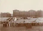 Uppställning med fanor och musikkår på Svea livgardes kaserngård vid firandet av Svea livgardes 400-års jubileum 1921. På gårdsplanen kung Gustaf V och i vagn drottning Victoria. Mellan husen i öster syns Göta livgardes exercishus och trädkronor i Göta Livgardes regementspark.