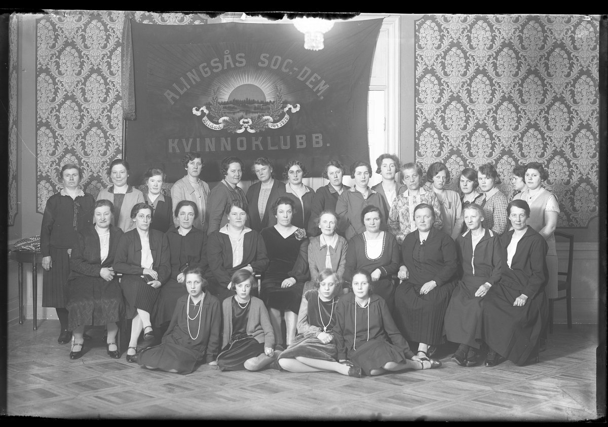 Gruppbild med Alingsås Socialdemokratiska kvinnoförening. 30 kvinnor i olika åldrar står uppradade framför föreningens fana. Fanan har texten "Alingsås Soc.-Dem. Kvinnoklubb. Bildad 18/2 1919" samt ekblad och en soluppgång.  Edith Gustafsson står som nummer två längst upp till vänster sett framifrån.