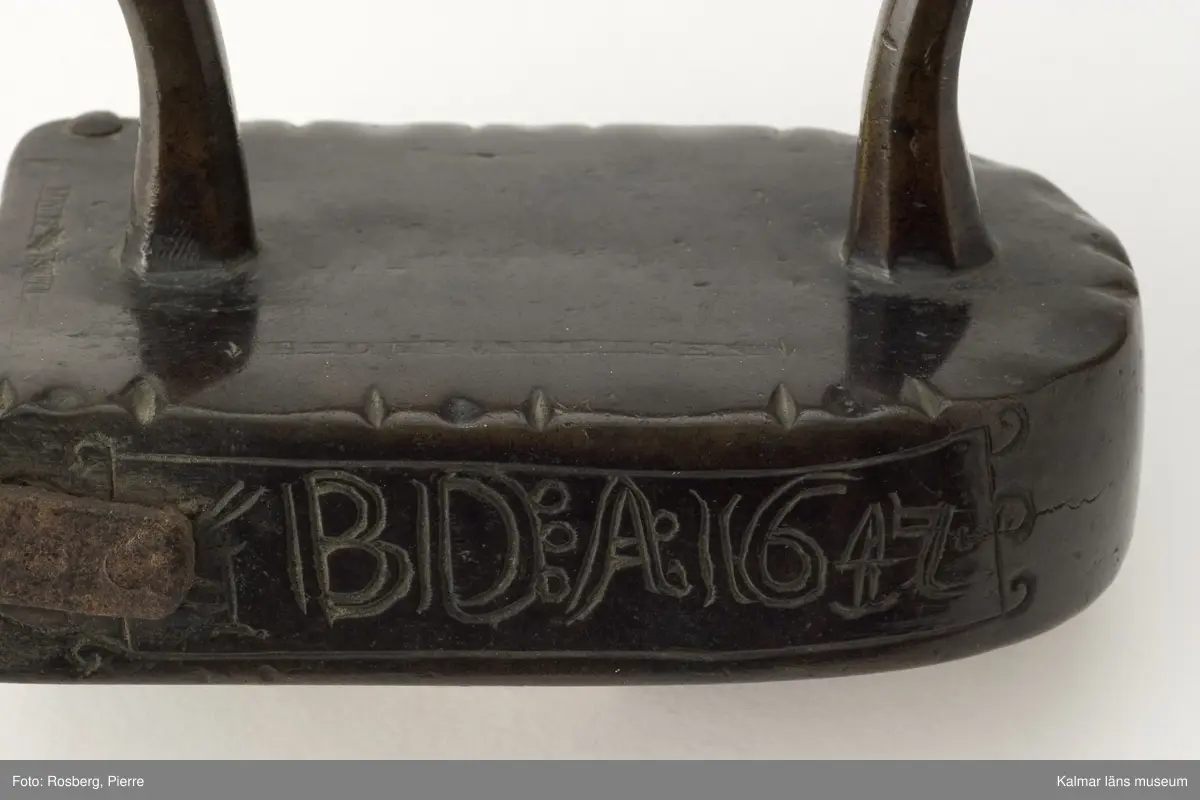 KLM 5926. Stykjärn. Av malm. Avsett för lod. Lucka baktill. Framtill rundat. Handtaget av malm. Text på smalsidan inom en ram: G.B.D.A:1647, på uppsidan inpunsat: PEDER NIELSEN. Luckan ornerad med halvmånformiga ornament,