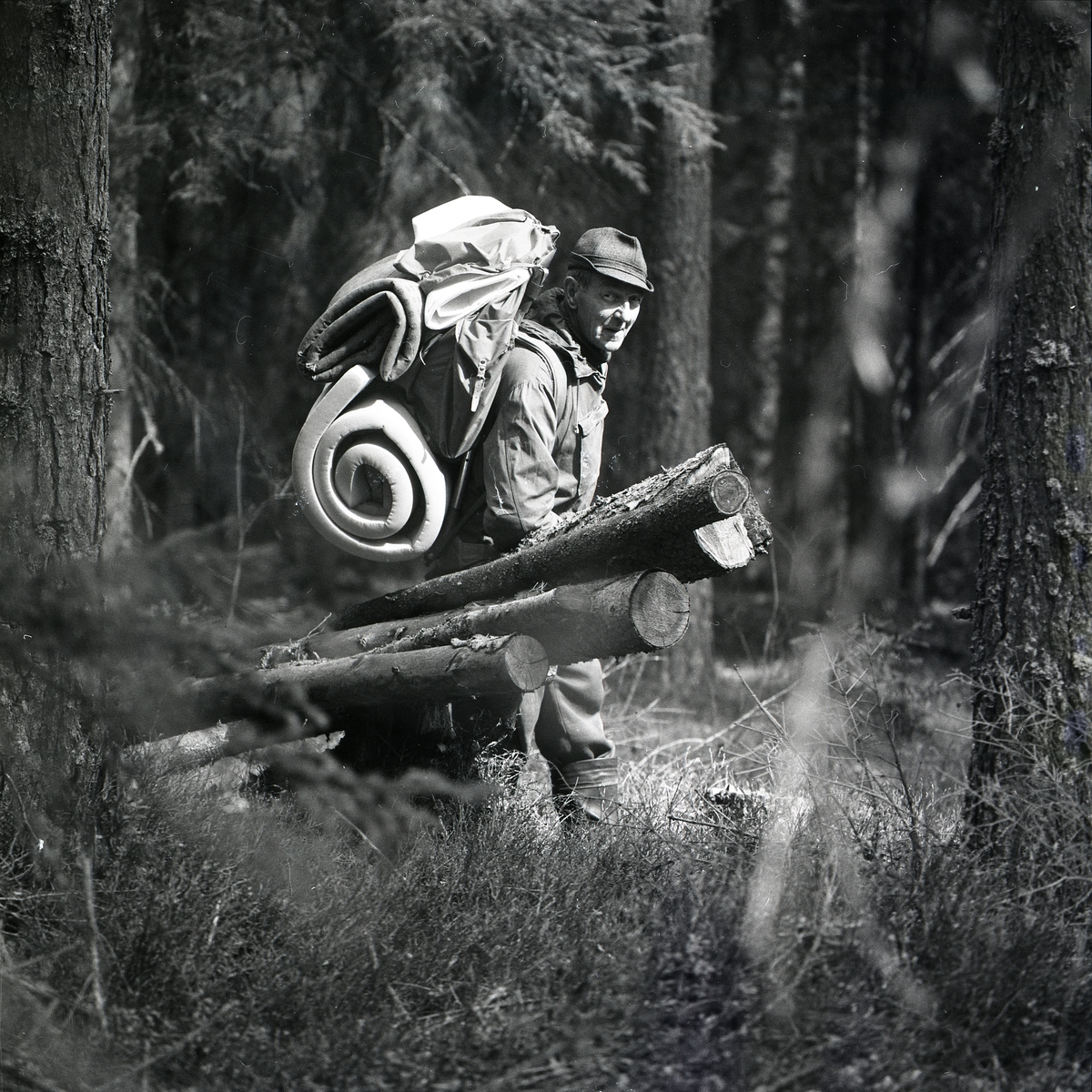 En man står vid några stockar i skogen våren 1975. Han bär en stor ryggsäck med mycket packning fäst på utsidan.